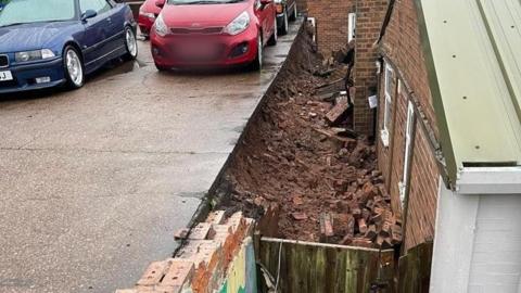 A collapsed wall between a car park and the back of a building at a lower level