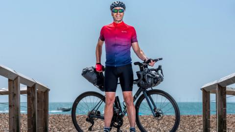 A man in cycling gear standing in front of his bike. He is on a beach with the sea behind him.