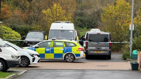 A police car parked in front of a police cordon. There are three cars behind the cordon