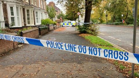 Blue and white tape saying "police line do not cross" with a police van and pavement in the background.