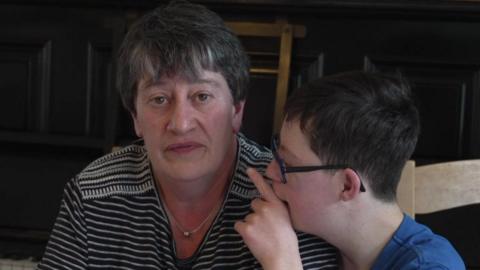 A woman with short brown hair wearing a black and white striped T shirt. Next to her sits a teenage boy with Down's syndrome wearing a blue T shirt and glasses.