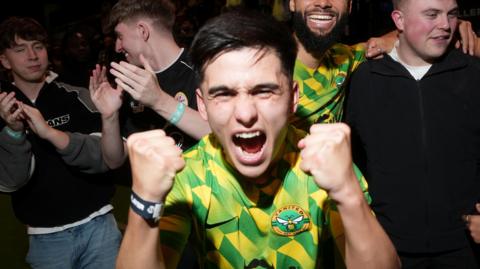 Bryan Ly wears a green and yellow Yanited jersey. He has his fists raised and clinched and is shouting into the camera. 