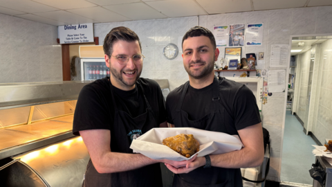 Two men with dark hair and beards in dark t-shirts holding a battered ball in white paper between them with silver counters and a white tiled wall behind them.