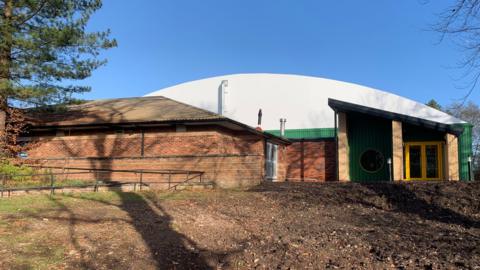 An indoor tennis centre building with a curved white roof and green and yellow entrance doorway. There is another low, brick building in front of it and rough ground in front of that. Shadows of trees are cast across the buildings.