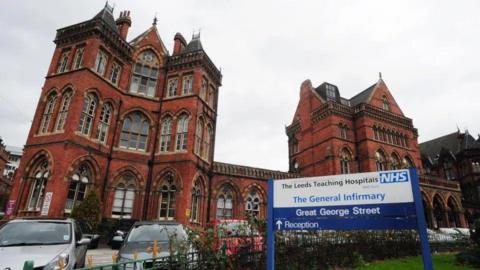 The outside of Leeds General Infirmary, a Victorian redbrick building with cars parked outside and a blue-and-white sign indicating it is a hospital.