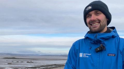 Joseph Earl, wearing a blue coat with Morecambe Bay Partnership logo and woolly hat, smiles. The salt marshes are in the background.