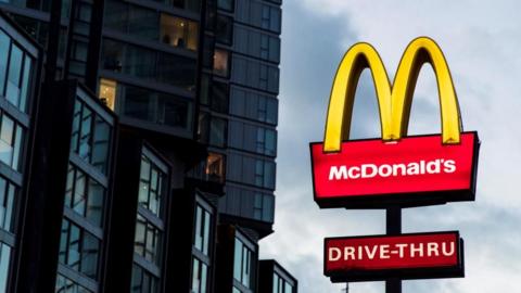 An illuminated McDonald's sign, consisting of a yellow M on a red block containing the word's McDonald's in white, is shown near a block of flats.