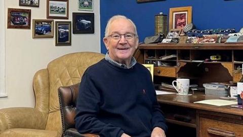 A man in a navy jumper, navy trousers and checked shirt. He has white hair and glasses and is smiling as he sits in a leather chair at a dark wood desk. The desk is piled with knick-knacks and family photos. Framed photos are mounted on a cream-painted wall to his left.