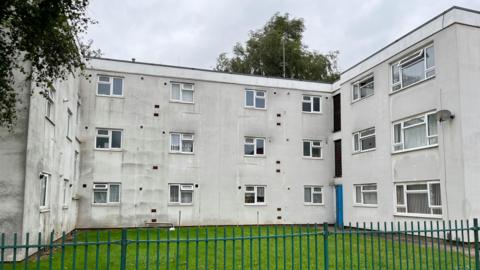 A three-storey block of flats. The building is off-white with multiple windows. In front is a green patch of grass