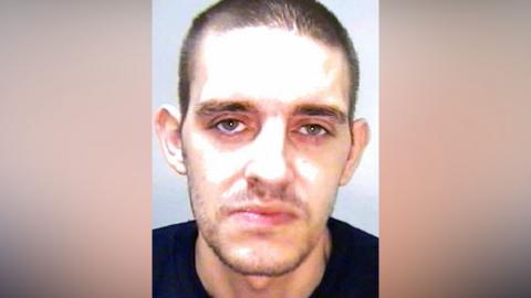 A police custody image of a man wearing a black t-shirt. He has a stubble beard and short dark hair and is staring expressionless into the camera.