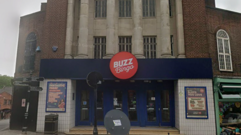 The front of an old cinema with large stone pillars on the first floor, and slim glass windows. the frontage has white tiles and blue doors and a sign. There is a red circle that reads "buzz bingo" in it