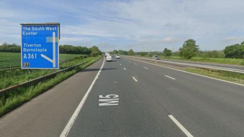 The M5 at junction 27 as the exit slip road starts with the blue motorway sign pointing towards Tiverton and Barnstaple A361 and the South West and Exeter on the left-hand side of the road.