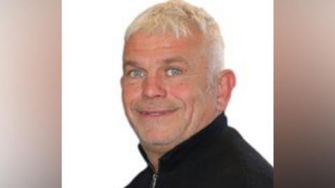A man with short white hair wearing a black jumper.