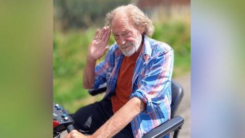 Mark Jackson, who is riding in his mobility scooter. He has his hand on his head in a salute to the camera, he is wearing a blue and orange large- checked shirt and an orange T-shirt. He has long grey hair and a grey beard.