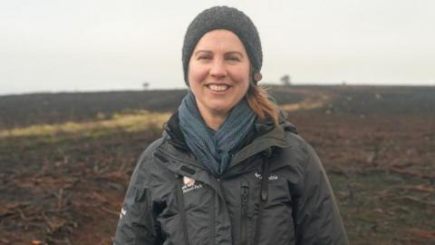 A woman, Elspeth, smiles at the camera. She is standing on moorland which is mostly blackened with some brown areas and a lighter brown line running through. She is wearing a hat and scarf, as well as a coat bearing the logo of the North York Moors National Park Authority.