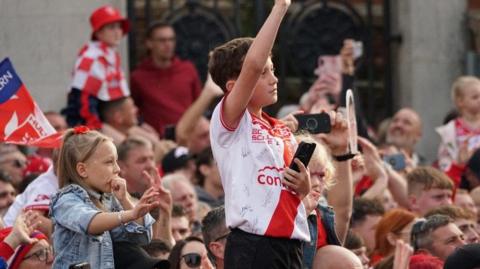 A young boy wearing a red and white Hull KR shirt is standing up with his arm in the air. A young girl is behind him in a jean jacket with her hand to her mouth.