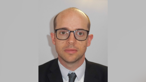A police head shot of a bald man wearing black-rimmed glasses. He is wearing a black suit, a white shirt and a grey tie. 