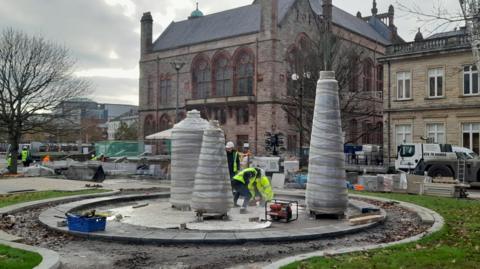 Workmen are erecting three oversized spools of cotton onto a stand at Derry's Harbour Square, part of a new piece of public art in the city remembering the factory workers of years gone by. The city's guildhall is seen in the background