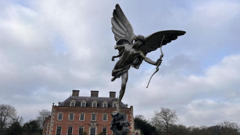 The replica Anteros statue at St Giles House. The winged god is holding a bow and arrow. In the background is the historic brick mansion topped with three large chimneys.
