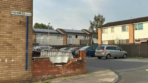 A brick wall with a sign on it reads 'Fisher Cl' which is next to a side street where a number of cars are parked. 