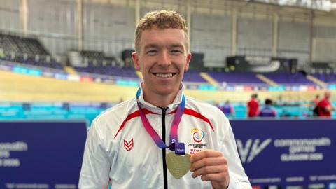 Matthew Bostock is standing in front of the velodrome, wearing a white sport jacket, and holding his gold medallion necklace. He has short light brown hair and is smiling.