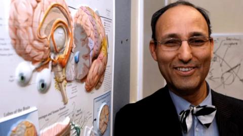 A man with glasses and bow tie stands next to a wall display of the human brain