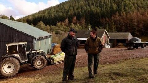 Laura and Mark Day, a couple in their 30s, are standing chatting in a farmyard dressed in dark trousers and jackets. He is wearing a cap and has a beard, she has short dark hair. 