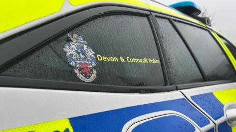 Close-up shot of a Devon and Cornwall Police car. It is mostly white, yellow and blue with a police emblem on a window.