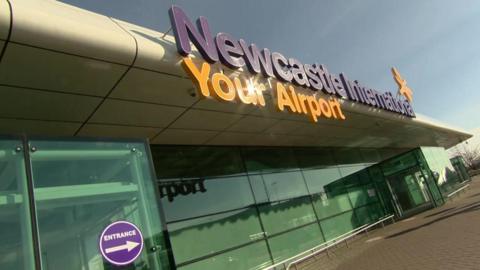 The entrance of Newcastle Airport. It is a glass building with Newcastle International Airport Your Airport sign in purple and yellow at the top, with a small plane next to it.