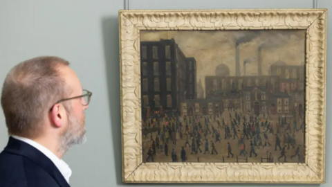 A man looks at Lowry's Going To The Mill as it hangs in a frame on a wall. It depicts an industrial Manchester landscape with matchstick figures walking in the foreground. 