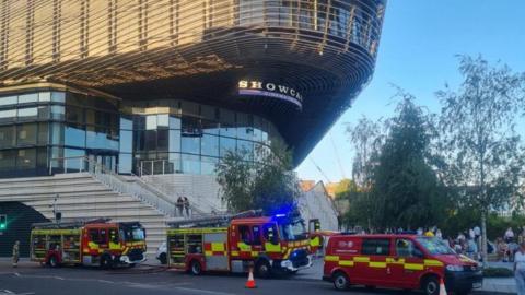 Fire engines and van parked up on a road outside a large shopping centre.