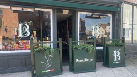 Benedictos in Worcester is pictured from the outside. The building is painted dark green with three green plant pots outside the building. Two of the large plant pots are slightly dented and scraped. 