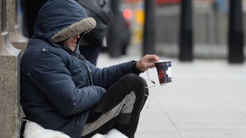 A man in a coat and tracksuit trousers holding up a coffee cup, asking for donations.