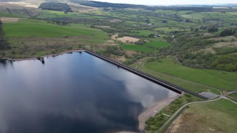 Drone image of Crai Reservoir. A large body of water can be seen in the foreground, surrounded by hills, fields and trees