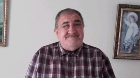 Paul Smith is smiling at the camera with a checked purple shirt buttoned up to his chin. He has light stubble and dark hair.