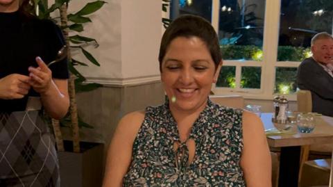 Sarbjit Budesha in a cafe or restaurant, sitting at a table in a floral shirt smiling while looking down