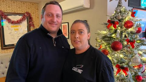 Man on the left and woman on the right, both in black hoodies, stood in front of a Christmas tree with red baubles.