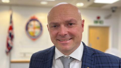 Andy Barnes has a shaved head and is wearing a navy suit jacket, white shirt and checkered tie. He is smiling inside the council chamber, where there is a union jack flag behind him.