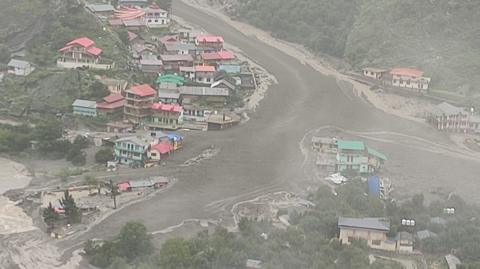 A still from a video showing the river of water flowing through the town. Brighly coloured houses can be seen on either side, as well as trees. 