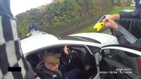 Daniel Pickavance arrest: a light-haired man sitting in the driver's seat of a pale car with his left hand raised. In the top right hand corner a hand is holding a yellow device which is pointed towards the car.