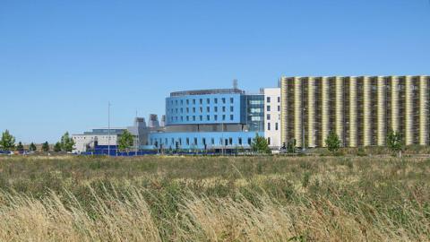 The Royal Papworth Hospital is part of the Cambridge Biomedical Campus. It is a large blue and cream-coloured building. In the foreground is a large field of long grass.