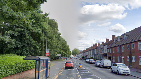 Cars drive along Newhampton Road East with other vehicles parked on the righthand side of the street outside a row of terraced houses.