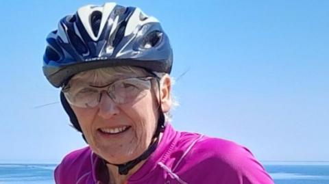 Pat Marshall, who is standing at the coast with her bicycle, is wearing a blue helmet, glasses and a pink top. She is looking at the camera and smiling slightly.