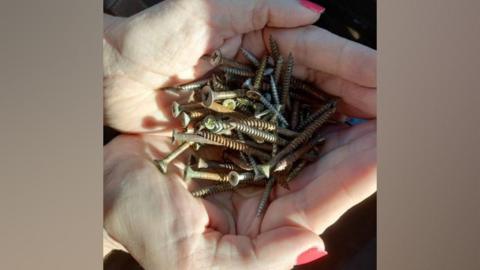 Two hands hold a small pile of rusty screws which were thrown onto roads in Whaplode. The female hands have nails which have been painted pink.