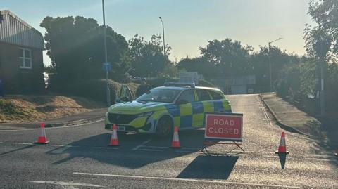 A police car parked in the middle of the road. There are cones blocking the junction with a red road closed sign.