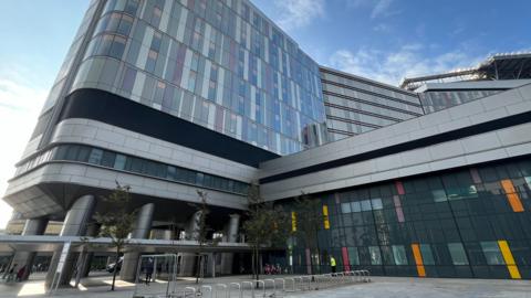 A view of the Queen Elizbeth University Hospital from the ground pointing up at the tall silver building, with patches of coloured glass on the outside of the building. The entrance has several silver columns around a courtyard