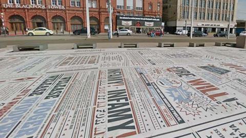 Street view image of the Comedy Carpet. A huge flat installation on the promenade full of text written in different fonts and sizes in red black and blue on a white background