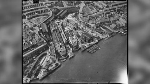 A black-and-white aerial shot of shipyards and the sea.