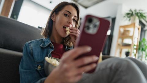 A woman eating popcorn from a bowl while scrolling on her red smartphone