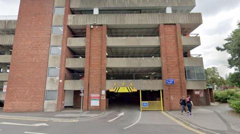 The front of a multi-storey car park, made up of red brick and grey concrete. It has five upper levels and the entrance has a blue sign saying 'IN'.